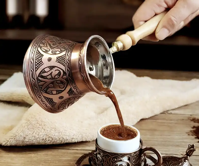 قهوة تصب من إبريق نحاسي مزخرف في فنجان على طاولة خشبية
